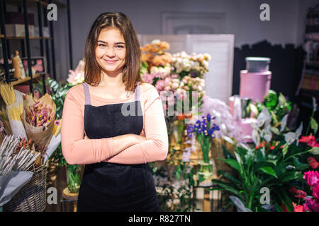 Bild von zuversichtlich oyung Blumen schauen auf Kamera. Sie stehen im Raum voller Blumen und Pflanzen. Junge Frau lächeln. Sie trägt schwarze Schürze. Stockfoto