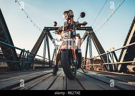 Close-up junge Frau sitzen auf dem Motorrad. Zwischenstopp auf dem neuen repariert vintage Holzbrücke. Biker Hintergrund. Outdoor extreme Aktivität. Wild road Abenteuer. Stockfoto