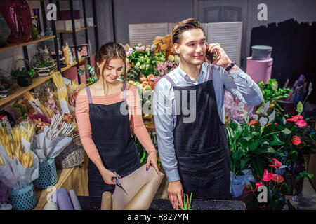 Junge Frau stand am Tisch und Schnitt von Papierrollen. Kerl stand hinter ihr und am Telefon sprechen. Er Lächeln. Sie sind im Raum voller Blumen und Pflanzen. Stockfoto