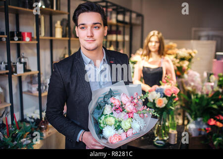 Stilvolle jungen Mann halten Schönen Blumenstrauß in den Händen und freuen uns. Er geht aus dem Blumenladen. Junge weibliche Floristen stehen hinter ihm. Stockfoto