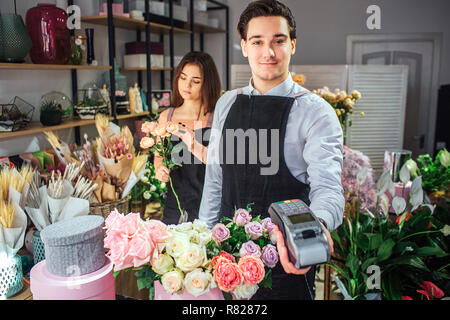 Schöner und fröhlicher junger Mann florist Suchen auf Kamera und halten Sie Geld therminal. Er stand am Tisch mit Blumen. Junge Frau hinter ihm steht, Blumenstrauß. Stockfoto