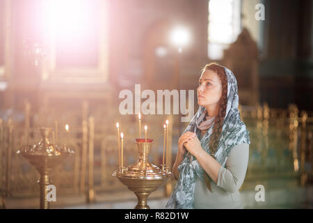 Russische schöne kaukasische Frau mit roten Haaren und einem Schal auf dem Kopf ist in der orthodoxen Kirche, leuchtet eine Kerze an und betet vor dem Symbol. Stockfoto