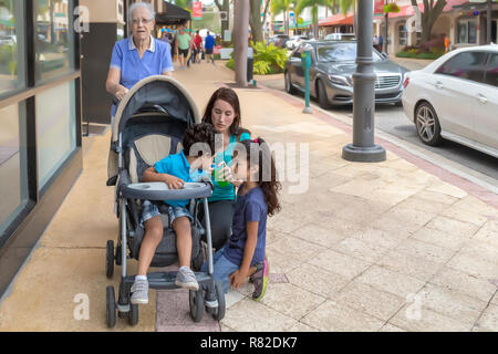 Familienausflug in der Innenstadt mit den Kindern. Die Großmutter hält den Kinderwagen, während die Mutter für eine sippy Cup für ihren kleinen Jungen und Tochter erreicht. Stockfoto