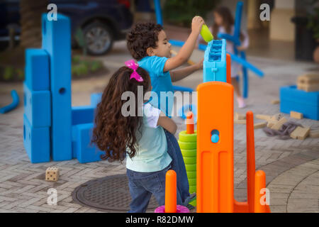 Die große Schwester hilft, ein jüngerer Bruder von ihm aufnehmen, während Er fügt die grünen Ringen in der Mega-Größe 4-Spiel, während andere Kinder spielen. Stockfoto