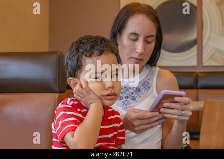 Ein gelangweilter kleiner Junge ist traurig, dass Mama hält texting. Eine Mutter an einem Kaffee Haus auf Ihrem Handy konzentriert, während das Kind hält seinen Kopf sitzen auf Mom. Stockfoto