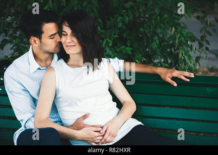 Eine junge moderne Paar sitzt auf einer Bank mit Blätter im Hintergrund, dass sie schwanger ist und Wiegen ihre runden Bauch sanft. Er umarmt sie zärtlich. Stockfoto