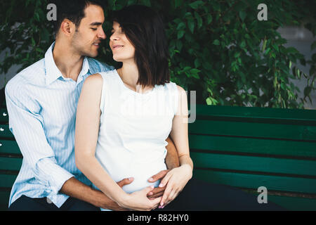 Eine junge moderne Paar sitzt auf einer Bank mit Blätter im Hintergrund, dass sie schwanger ist und Wiegen ihre runden Bauch sanft. Er umarmt sie zärtlich. Stockfoto