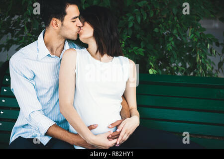 Eine junge moderne Paar sitzt auf einer Bank mit Blätter im Hintergrund, dass sie schwanger ist und Wiegen ihre runden Bauch sanft. Er umarmt sie zärtlich. Stockfoto