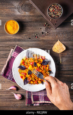 Mann essen Herbst Salat mit Kürbis, Karotte, Tomate und Basilikum auf dem hölzernen Tisch mit Gewürzen Stockfoto