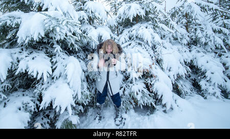 Eine Frau mittleren Alters mit blonden Haaren spielt mit Schnee. Sie ist glücklich und schüttelt ihre kalte Hand. Im Hintergrund sind schneebedeckte Tannen. Stockfoto