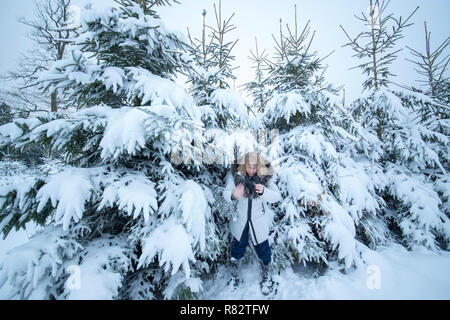 Eine Frau mittleren Alters mit blonden Haaren spielt mit Schnee. Sie ist glücklich und schüttelt ihre kalte Hand. Im Hintergrund sind schneebedeckte Tannen. Stockfoto