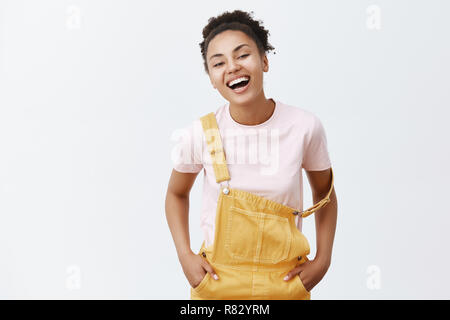 Charmante Freundin mit dunkler Haut und lockiges Haar, laut lachen, tragen Süße gelbe Overalls mit Gurt fallen auf den Arm, halten sich an den Händen in der Tasche und freudig lächelnd, während im Gespräch mit Freund Stockfoto