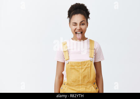 Die Jugend ist für Party und Chillen. Portrait von Sorglos glücklich Afrikanisch-amerikanische Frau im gelben Overalls über trendy t-shirt, die Zunge und freudig Winking, verbringt den Sommer mit Freunden Stockfoto
