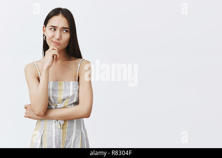 Hmm, lass mich nachdenken. Portrait von zweifelhafter gut aussehenden asiatische Frau in passendem trendy Outfit, grinsend und den Blick nach rechts, während die Hand am Kinn, Denken, zögern über grauer Hintergrund Stockfoto