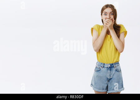 Schockiert beeindruckt attraktive kaukasische Mädchen im gelben T-Shirt und Shorts keuchend über Mundpropaganda von Schock und Überreaktion mit Palmen an Kamera starrte gegen grauen Hintergrund erstaunt Stockfoto