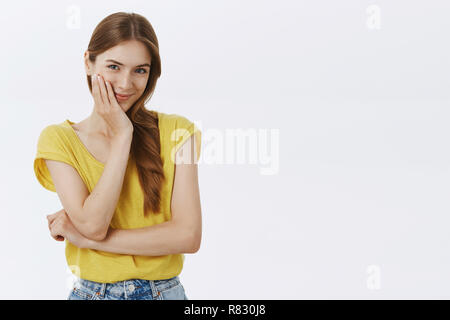 Taille-up Shot der netten weiblichen Freundin in Gelb - thisrt berühren sie sanft in der Wange lächelnd sinnlich und Blick auf die Kamera mit Zuneigung und Liebe über den grauen Hintergrund posiert im gelben T-Shirt Stockfoto