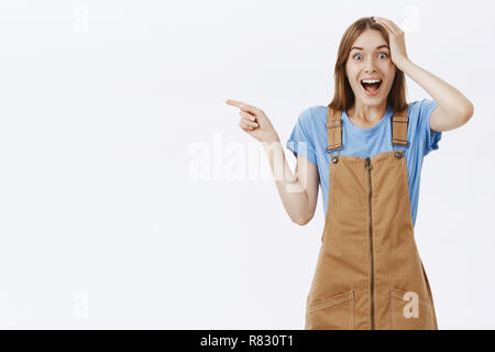 Portrait von überrascht gut - beeindruckt auf der Suche kaukasischen Frauen in braunen Overalls Hand am Kopf und nach links Gefühl begeistert und überrascht seing awesome Person lebendig über graue Wand Stockfoto