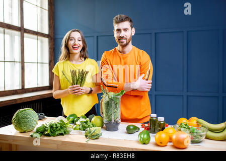 Bunte Porträt eines jungen Paares in helle Kleidung Smoothie zu machen auf dem Tisch voller grünes Gemüse auf dem blauen Hintergrund im Innenbereich Stockfoto