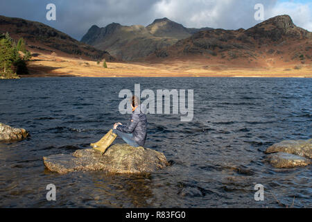 Frau sitzt auf einem Felsen in der blea Tarn bewundern Sie die Landschaft, die die Langdale Hügel, Cumbria, Großbritannien. Stockfoto