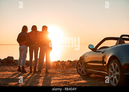 Fröhlicher junger drei Frauen, die in der Nähe von Cabriolet wathing bei Sonnenuntergang Stockfoto