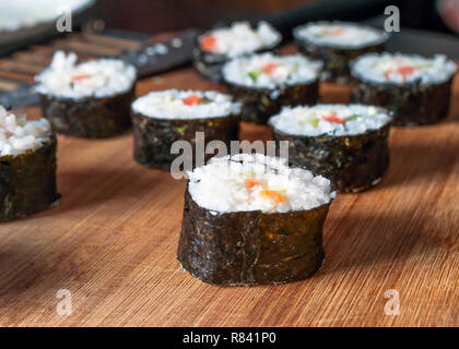 Chumaki Sushi Rollen mit Thunfisch, Lachs, Reis, Gurken, Avocado und Nori Algen auf einem Holzbrett mit Küchenutensilien Unscharf im Hintergrund. Stockfoto