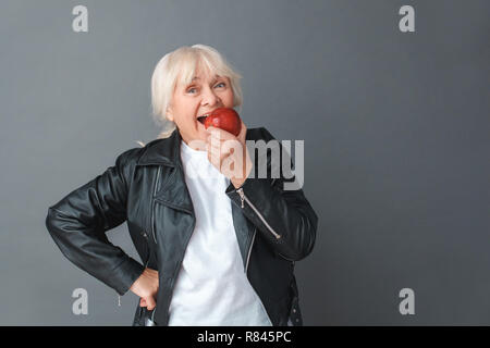 Ältere Frau in Leder Jacke Studio stehen isoliert auf Grau essen Apple freudige Stockfoto