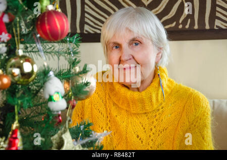 Porträt der alten Großmutter zu Hause in das neue Jahr. Kaukasische positiv, freundlich Granny in festlichen, weiß gestrickte Pullover. Ältere gemütliche Frau in der Nähe von Weihnachten, Baum. ältere Dame schaut in die Kamera. Stockfoto