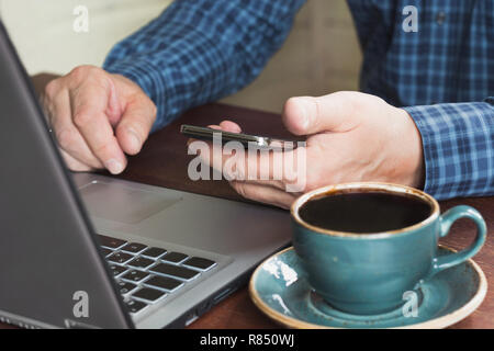 Seitenansicht schoss der Hände eines Mannes mit Smartphone und Laptop an den hölzernen Tisch sitzen bei einer Tasse Kaffee. Close Up. Stockfoto