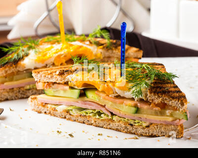 Club Sandwich mit Ei, Gurke, Tomate, Schinken. Zwei Club Sandwiches auf weiße Platte auf dunkelbraunen hölzernen Tisch im Restaurant Stockfoto