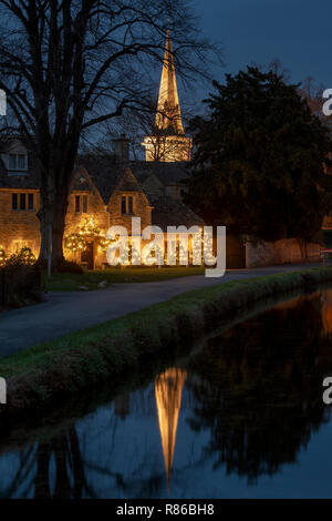 Weihnachtslichter auf einem cotswold-Häuschen mit der Kirche spiegeln sich in Lower Slaughter in der Nacht im Flussauge wider. Cotswolds, Gloucestershire, England Stockfoto