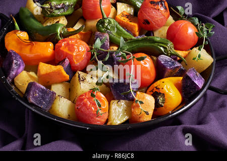 Rustikal, Ofen gebackenes Gemüse in Auflaufform. Saisonale vegetarische vegane Mahlzeit auf dunkel lila Bettwäsche Handtuch. Zutaten: Kartoffeln, Lila und süßen potat Stockfoto