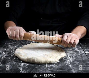 Koch in einem schwarze Tunika, rollt Teig für Pizza auf einem Schwarzen Tisch mit einem hölzernen Rolling Pin Stockfoto
