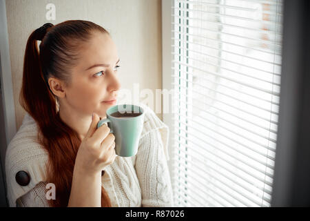 Tee trinken, sitzen auf dem Fenster der Kaukasischen schöne Frau mittleren Alters mit roten Haaren Stockfoto