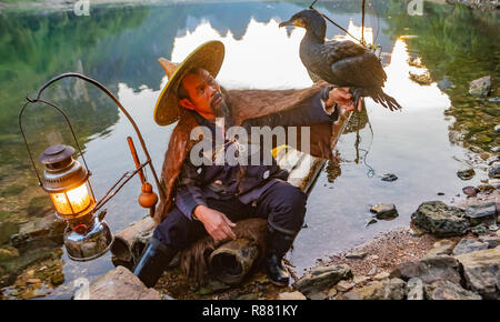 Chinesische Kormoran Fischer holding Kormoran Vogel auf dem Arm auf dem Floß in See in Guilin, China. Fischer tragen Stroh Hut, Umhang und hat Laterne auf floß. Stockfoto