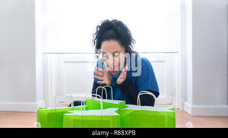 Portrait von normalen mittleren Alter Frau mit blauen Augen, weiße Haut und schwarze Haare macht eine lustige Überraschung Ausdruck bei vielen grünen Tüten voller Weihnachtsgeschenke von Winter Sales Stockfoto