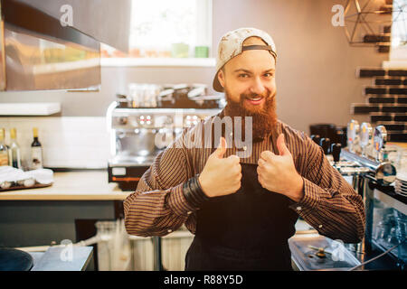 Glückliche junge Barista stehen und großen Daumen oben auf beiden Händen. Er Blick auf Kameras. Kerl stand in der Küche. Licht kommt aus Fenster Stockfoto