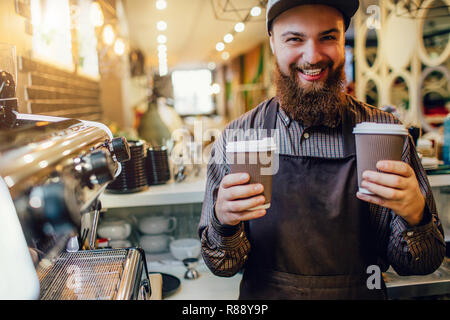 Happy bearded Barista zwei Tassen auf einen anderen Betrieb. Er lächelt auf Kamera. Kerl stehen in der Küche in der Nähe der Kaffeemaschine Stockfoto