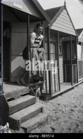 1930er Jahre, ein englischer Mann in ein Streifiges einteiligen sleeveless Badeanzug der Ära, stehend auf der Veranda einer Strandhütte in Southend-on-Sea, England, ein beliebter Ferienort für Menschen in Essex und messetermin London in dieser Zeit leben. Stockfoto