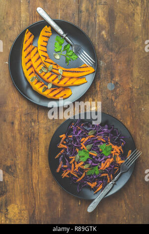 Gegrillte geschnittenen Kürbis mit Petersilie und Kürbiskerne, Salat von lila Kohl, Karotten und Samen in schwarzen Keramikplatten auf dunklen alten hölzernen Oberfläche, Stockfoto