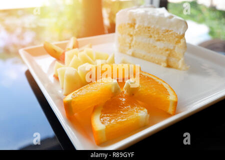Stück Obst und Milch Kuchen auf weiße Platte/Nahaufnahme von frischen Orange Traube und Apfel Obst Slice auf Tablett mit Kuchen sahne Vanille Dessert auf den Tisch Stockfoto
