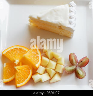 Stück Obst und Milch Kuchen auf weiße Platte/Nahaufnahme von frischen Orange Traube und Apfel Obst Slice auf Tablett mit Kuchen sahne Vanille Dessert auf den Tisch Stockfoto