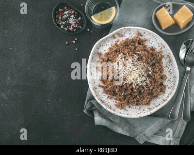 Buchweizen Risotto mit getrockneten Pilzen Parmesan in Handwerk Platte auf schwarzem Zement Hintergrund. Glutenfreie und vegetarische Buchweizen Rezept Ideen. Kopieren Sie Platz. Ansicht von oben oder flach-legen. Getonten Bild Stockfoto