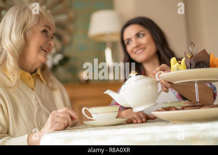 Verschwommen Brunette Tochter gießt Tee aus Weisse Teekanne zu Ihrem älteren Schöne Mutter im Cafe. Stockfoto