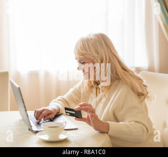Online einkaufen. Elegante reife Frau Typen auf dem Laptop, während Sie die Kreditkarte. Tasse Kaffee steht in der Nähe der Laptop, auf dem eine Frau mit einem Banki Stockfoto