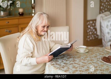 Horizontale Schoß der schönen alten Frau über das Buch auf den Tisch, während Sie Kaffee und Kekse. Stockfoto