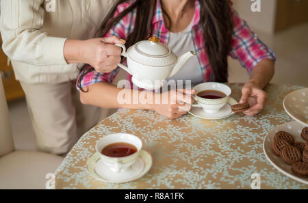 7/8 Schuß von Frau Gießen schwarzer Tee in Tochter s Cup. Frau hält eine leckere Cookies Während Ihre Tasse mit aromatischen Kaffee gefüllt wird. Stockfoto
