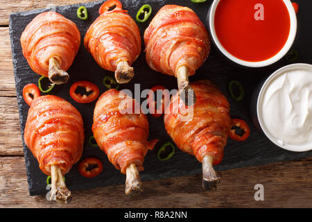 Würzige Lutscher gebratene Hähnchenschenkel in Speck mit Soßen serviert schließen eingepackten auf einer Schiefertafel Board auf dem Tisch. horizontal oben Ansicht von oben Stockfoto