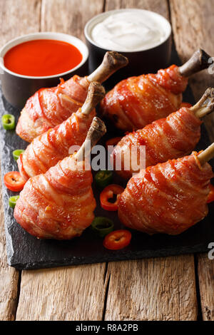 Würzige Lutscher gebratene Hähnchenschenkel in Speck mit Soßen serviert schließen eingepackten auf einer Schiefertafel Board auf dem Tisch. Vertikale Stockfoto