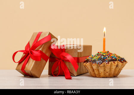 Lecker geburtstag Muffin mit Schokoladenüberzug und Karamell, mit brennenden festliche Kerze und Geschenkboxen auf beigen Hintergrund eingerichtet. Happy birthday Minderen Stockfoto