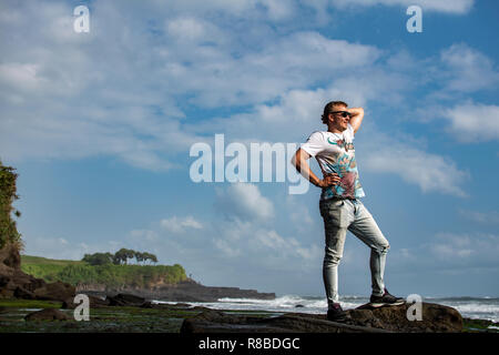 Junger Mann in t-Shirt am Winter-Strand bei Sonnenuntergang. Wolken bei Sonnenuntergang Himmelshintergrund und Felsen sind von hinten sichtbar. Hinten Sie Ansicht von. Stockfoto